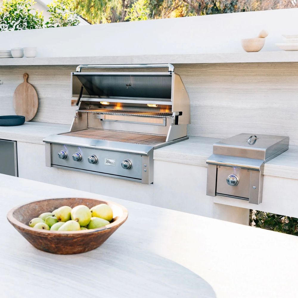 A stainless steel outdoor gas grill with a lid is mounted on a light stone kitchen island, with a matching side storage compartment nearby. The scene is set in a bright, modern outdoor kitchen with a bowl of green fruit in the foreground.