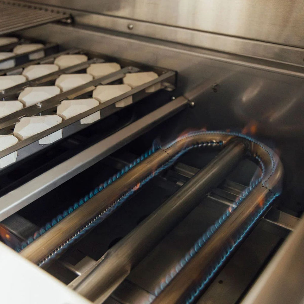 Close-up of a stainless steel gas grill or oven interior with blue gas flames burning along curved metal tubes and white ceramic racks visible in the background.
