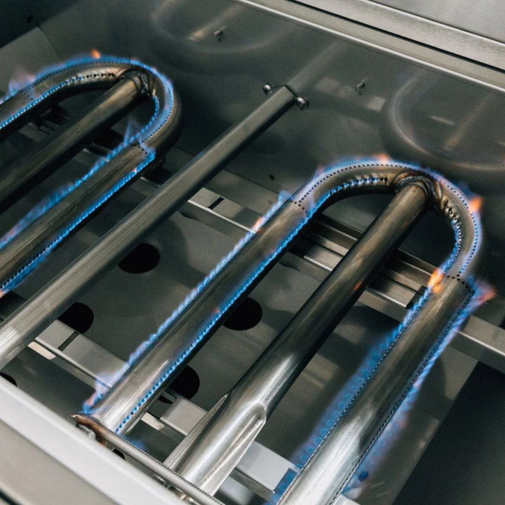Close-up of a stainless steel gas grill burner with blue flames ignited along the curved metal tubes inside the cooking chamber.