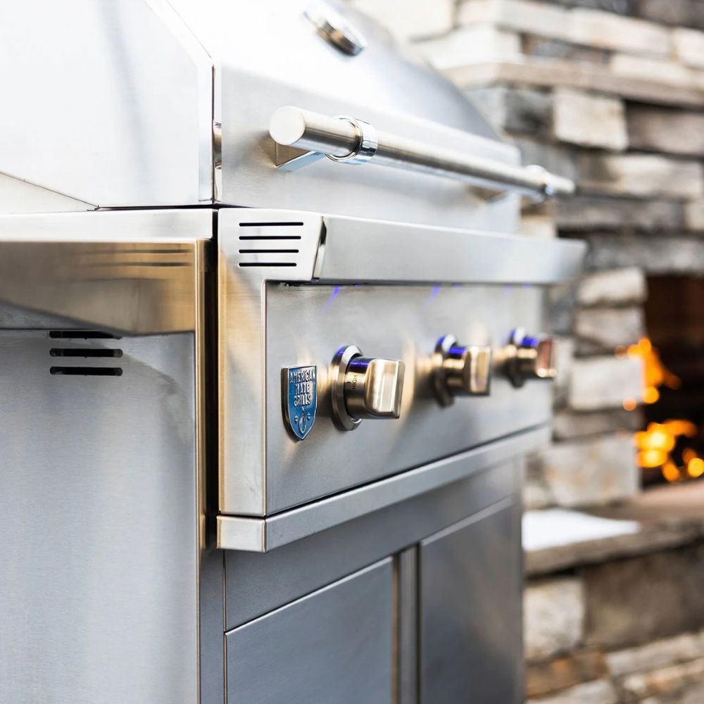 A close-up of a stainless steel outdoor gas grill with four control knobs and a blue logo badge, set against a blurred stone wall and fire.