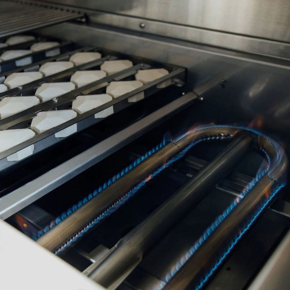 Close-up of a gas grill or oven interior with blue gas flames burning along metal tubes and white ceramic racks holding food.