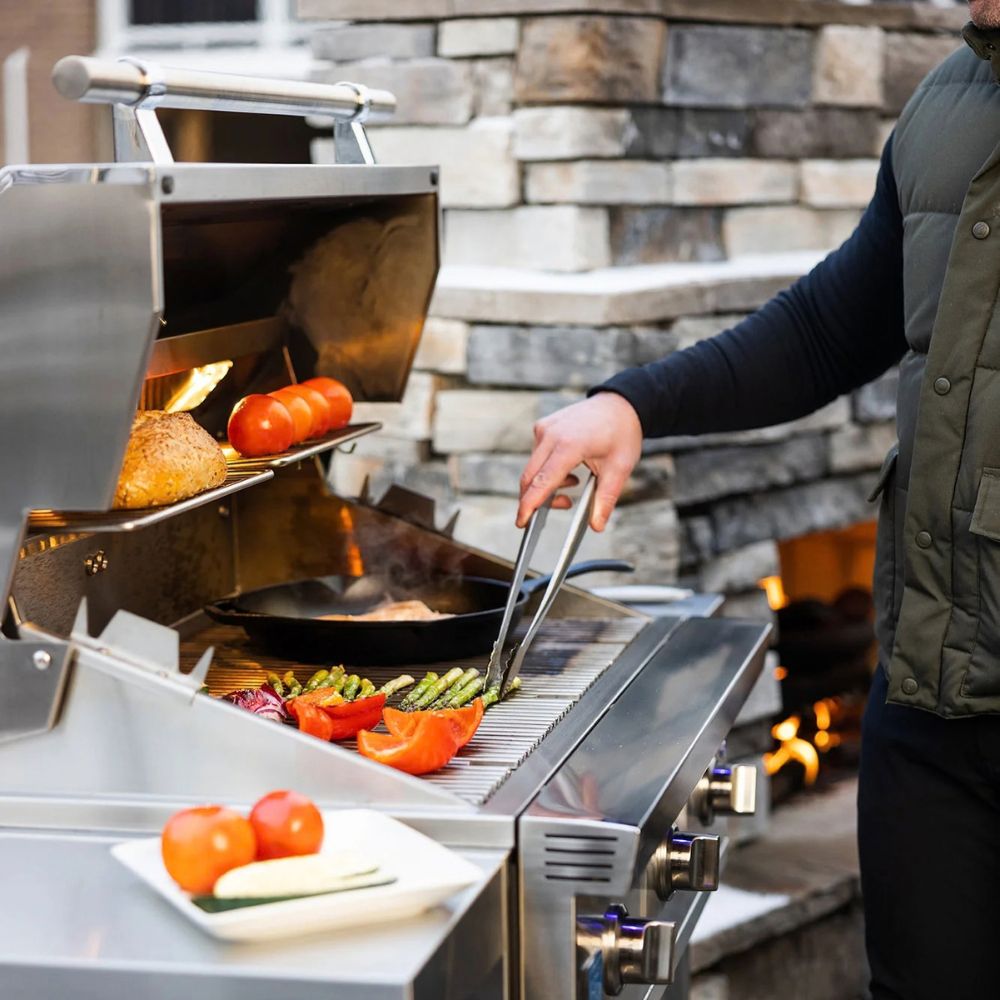 A stainless steel outdoor gas grill in use, with a person using tongs to turn food on the grates. Tomatoes, asparagus, and bread are visible on the grill, with a stone wall and fire pit in the background.