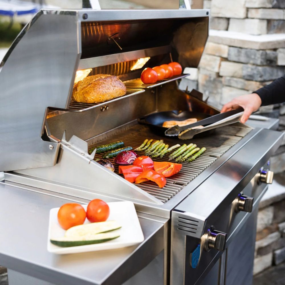 A stainless steel outdoor gas grill is shown with food cooking on the grates and interior shelves. A person's hand holds tongs over the grill, and a plate with tomatoes and zucchini sits on the grill's side.