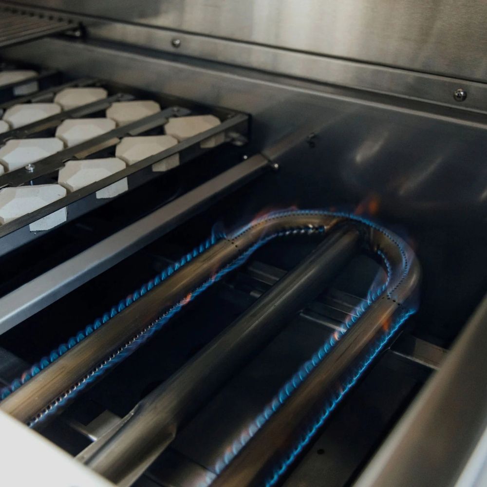 Close-up of a stainless steel gas grill interior with two blue-burning gas jets and white ceramic burner elements on a rack.