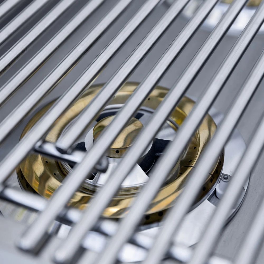 Close-up of a stainless steel grill grate overlaying a brass burner assembly on a metal grill surface.