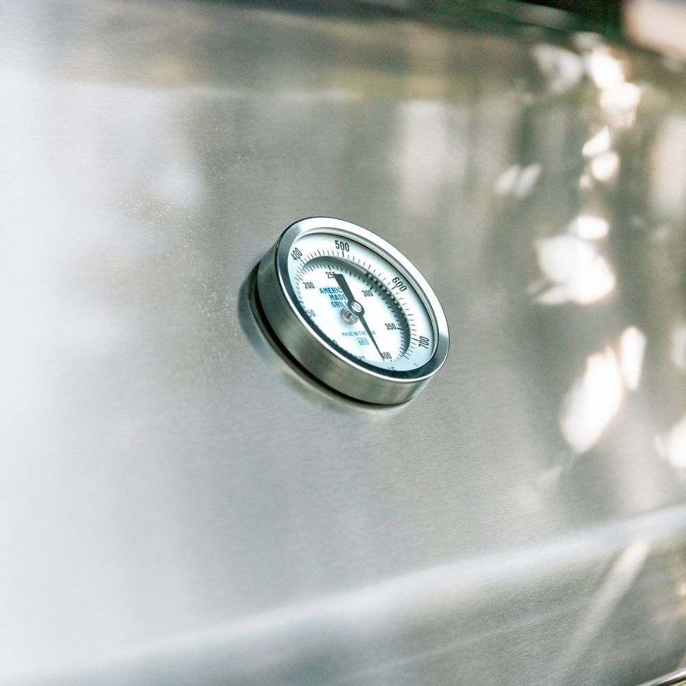 A round stainless steel temperature gauge mounted on a brushed metal surface, photographed close up with soft natural light and blurred background reflections.