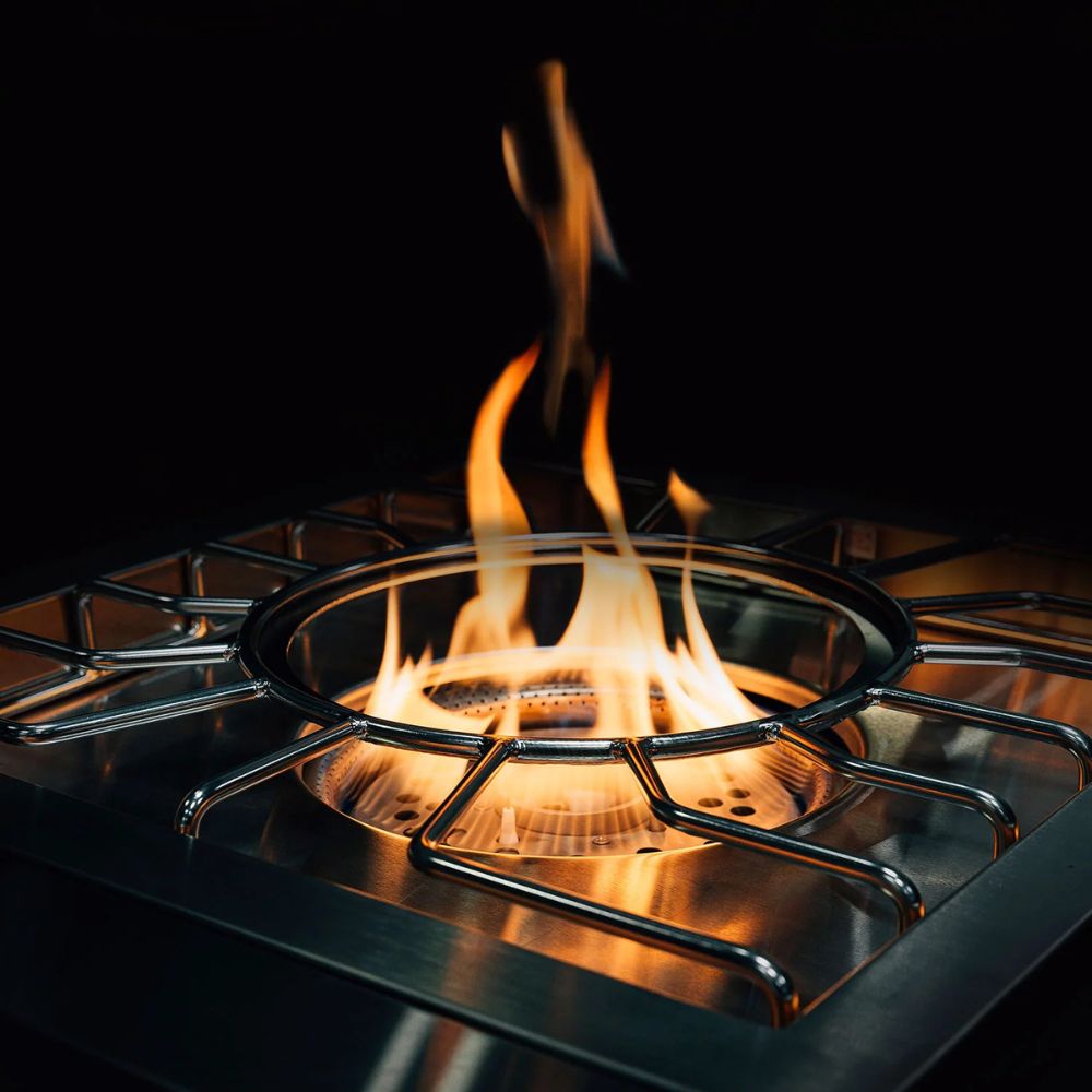 A close-up of a stainless steel tabletop fireplace or brazier with a circular fire burning beneath a metal grate, set against a dark black background.