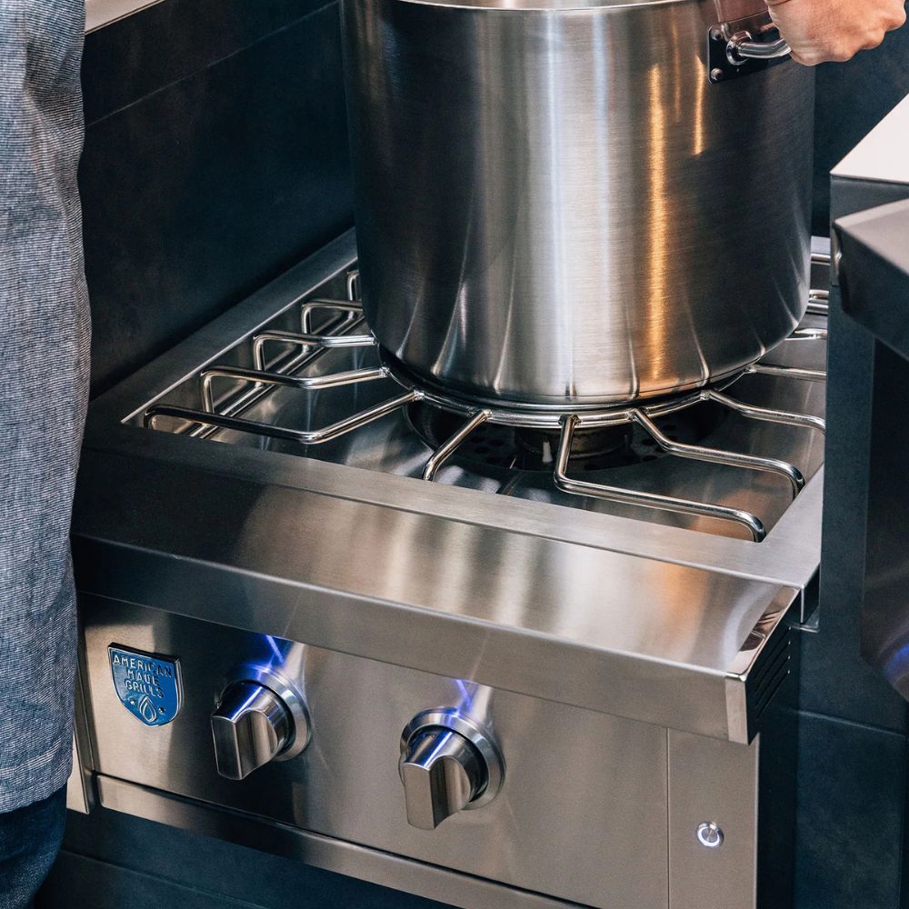 A stainless steel commercial gas range with two front control knobs and a large pot on a burner, photographed in a kitchen setting with part of a person visible at the left edge and right edge.