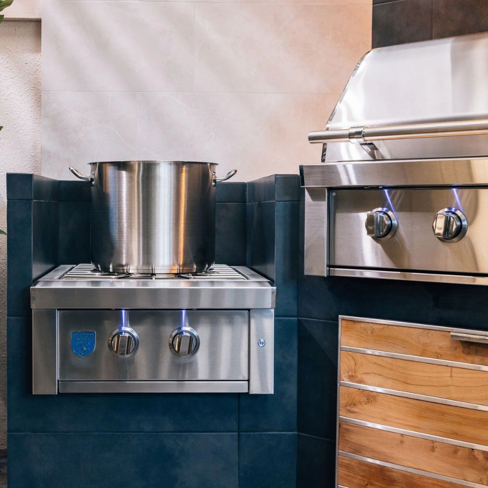 A stainless steel outdoor gas cooktop with blue flames on a dark tiled base, shown next to a larger stainless steel grill with wood-grain cabinets.
