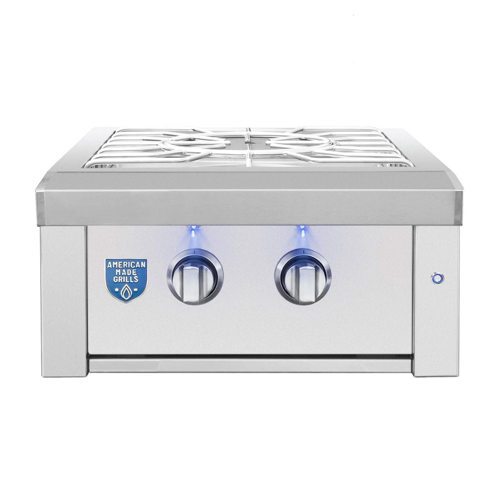 A stainless steel outdoor gas grill with two control knobs, blue indicator lights, a white grate on top, and an American Made Plus badge on the front, shown against a plain white background.