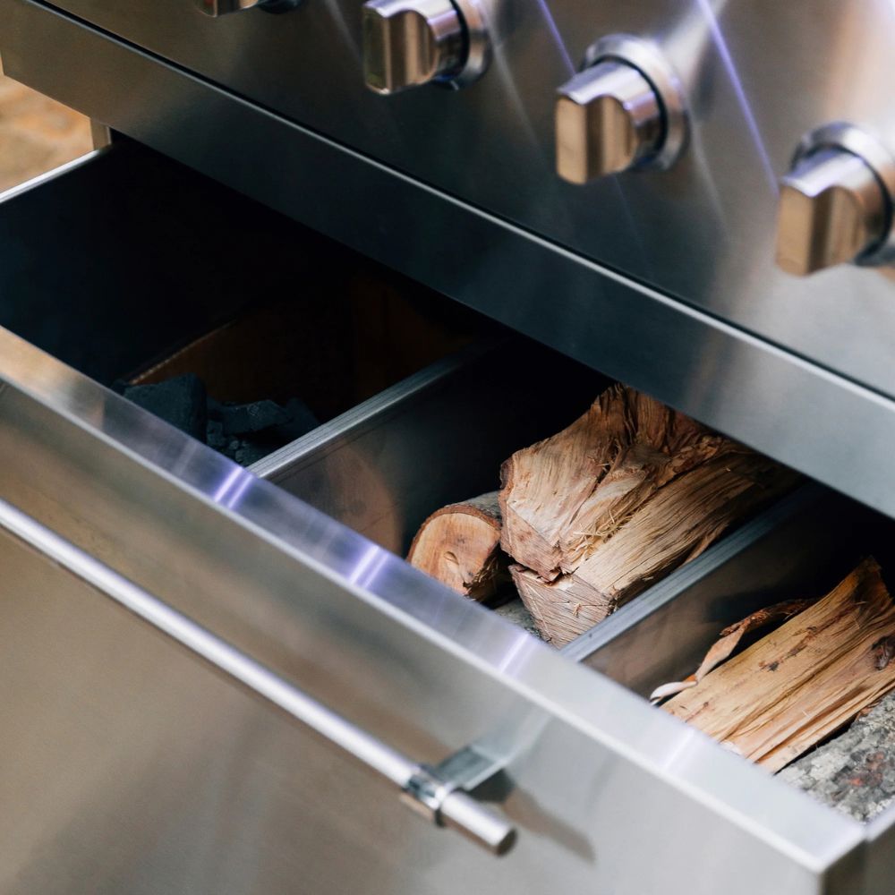 Close-up of a stainless steel outdoor grill drawer open, showing wood logs and coals inside, with part of the control knobs visible at the top.