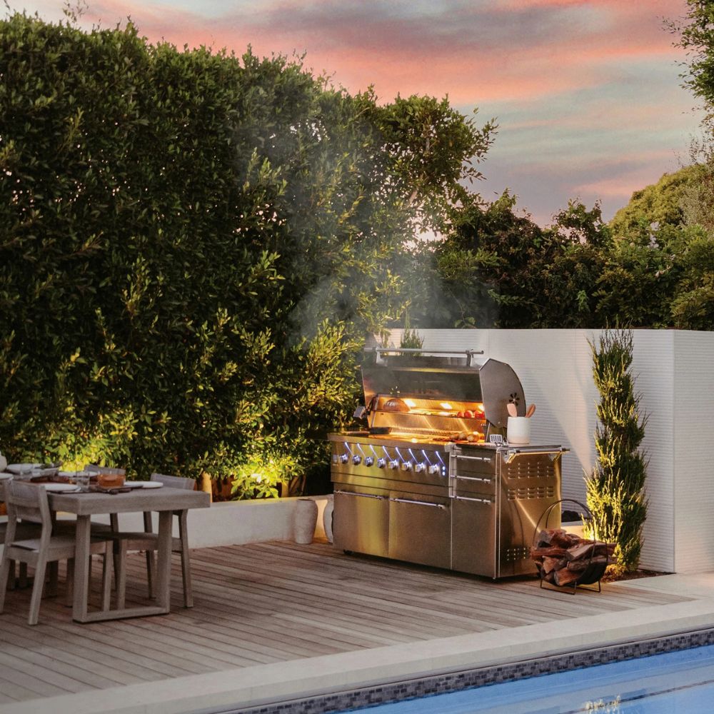Outdoor kitchen with a grill on a wooden deck by a pool at sunset.