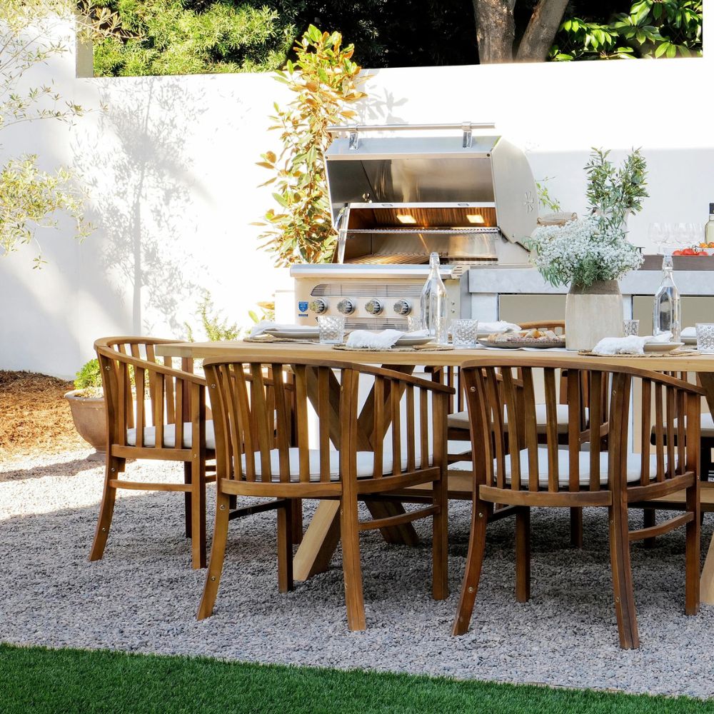 Outdoor dining setup with a rectangular wooden table and four matching slatted chairs on a gravel patio, with a stainless steel grill and kitchen island in the background against a white wall.