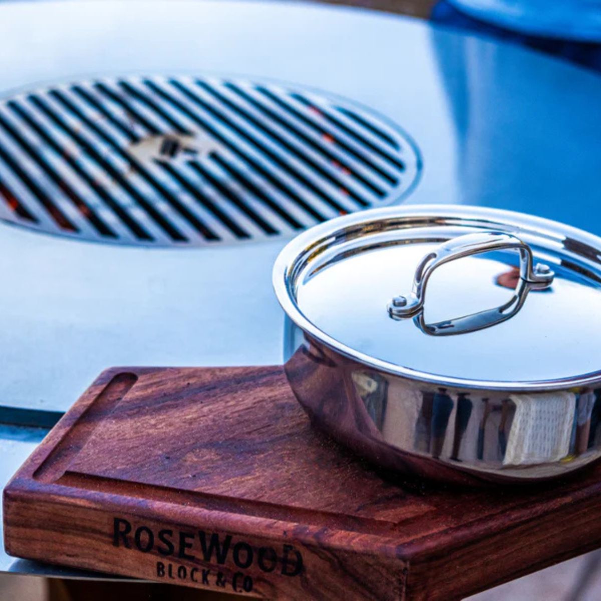 Stainless steel pot with lid on a wooden block labeled 'Rosewood Block & Co' on a blurred background