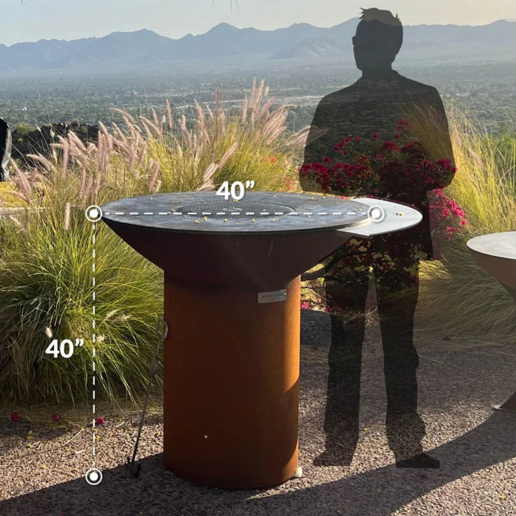 Outdoor setting with a large round table and a person standing nearby, with mountains in the background.