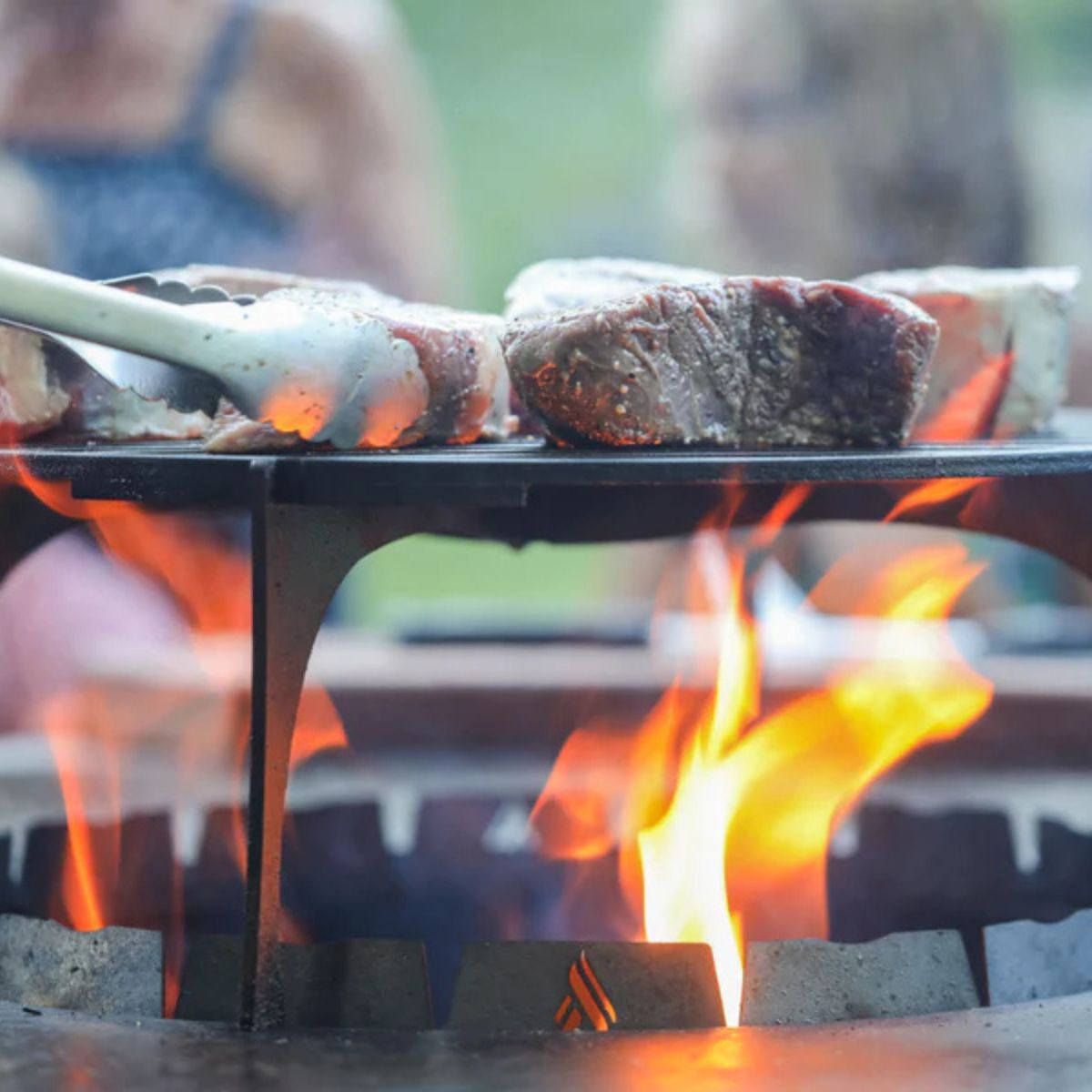 Steaks being cooked on a grill with flames and people in the background