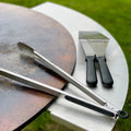 Barbecue tools including tongs and a scraper on a metal surface with grass in the background.