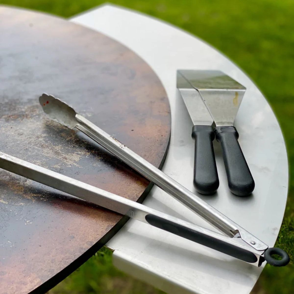 Barbecue tools including tongs and a scraper on a metal surface with grass in the background.