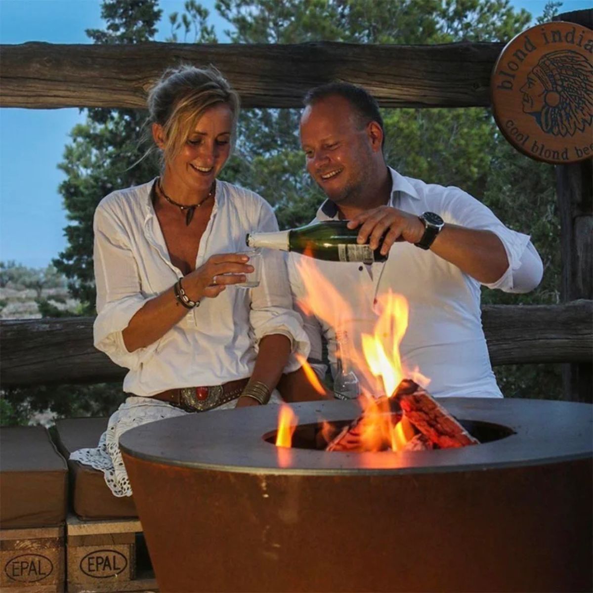 Two people by a fire pit, one pouring champagne, with a scenic background.