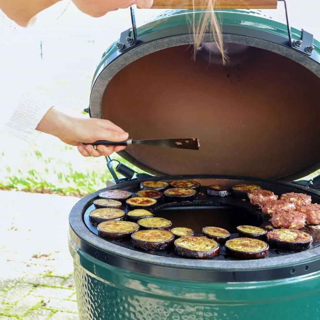 Person grilling vegetables and meat on a large green grill outdoors.